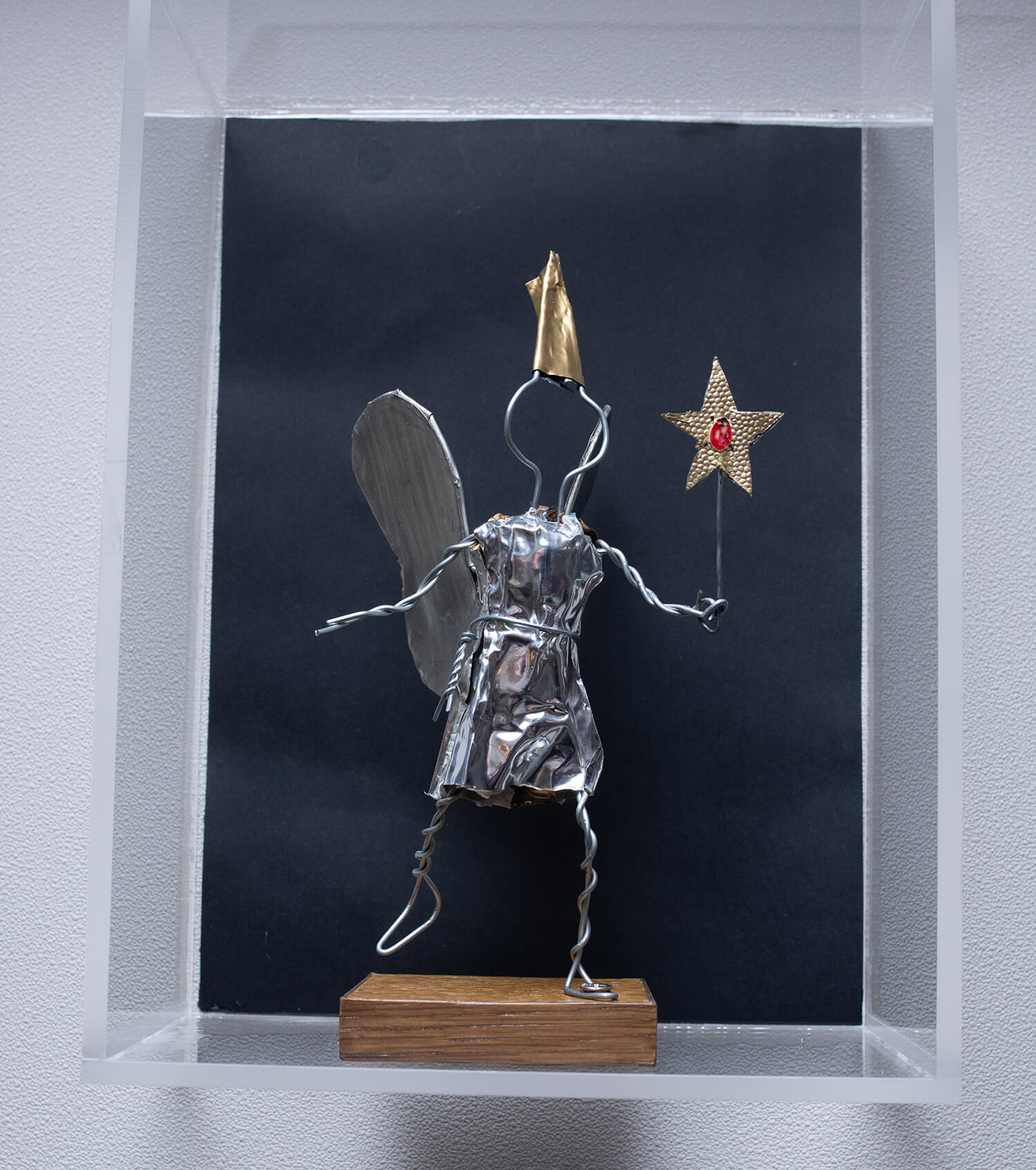 We see the small figurine of the Bulléo Fairy on a rectangular wooden base, made from the twisted wire of the muselet and the silver collar of the champagne cork. She is holding a magic wand in the shape of a golden star with a ladybug in the center. She has silver wings and a golden pointy hat.