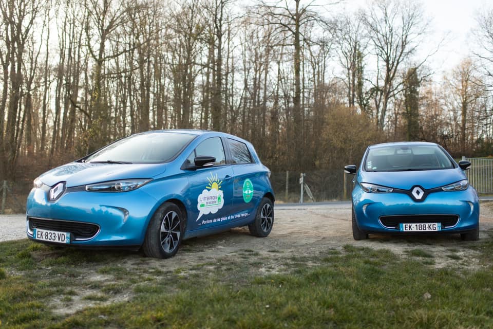 We see the 2 electric Renault Zoé parked on a dirt road in front of trees on the side of a road