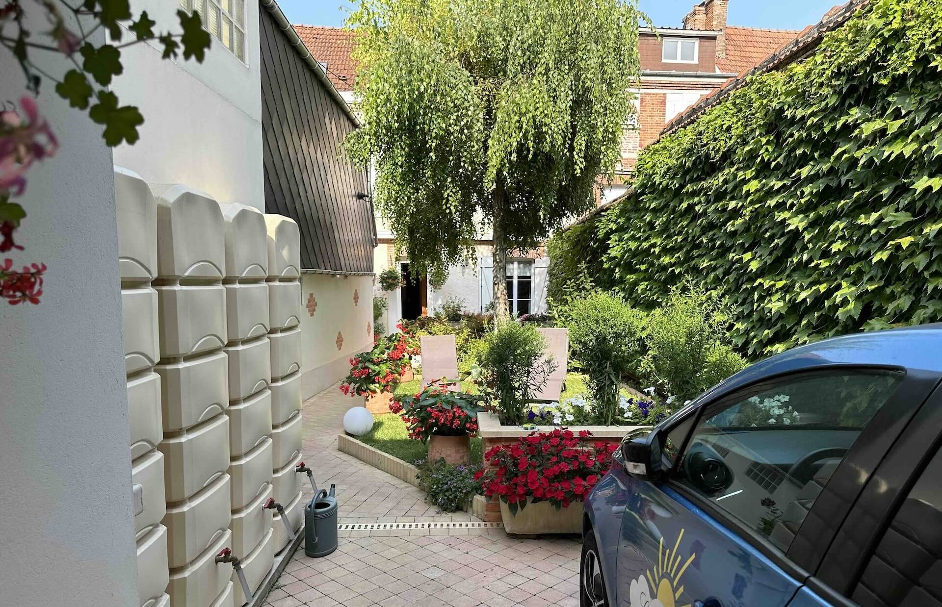 We have a view of the Linterfaces garden from the entrance courtyard. There is vegetation on the right wall, flowers, a tree, a path leading to the house at the far end, water collectors on the left and the front of one of the electric cars at the bottom right.
