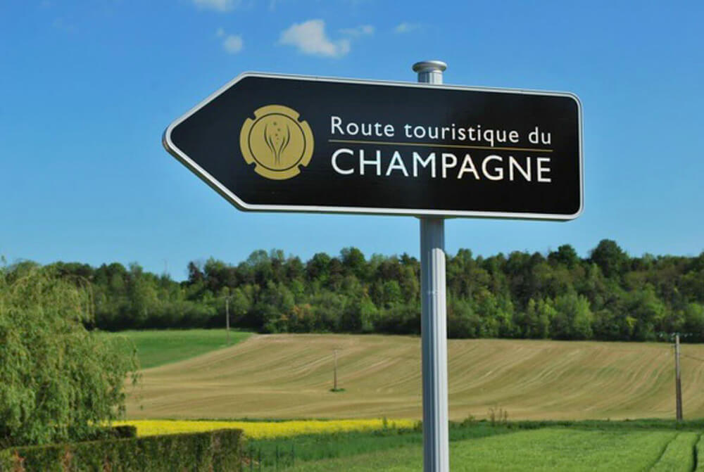 In the foreground we see a black arrowed sign indicating “Champagne tourist route” with agricultural fields and large trees in the background.