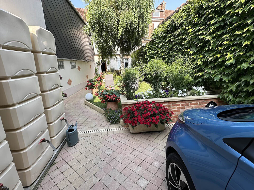 We have a view of the Linterfaces garden from the entrance courtyard. There is vegetation on the right wall, flowers, a tree, a path leading to the house at the far end, water collectors on the left and the front of one of the electric cars at the bottom right.