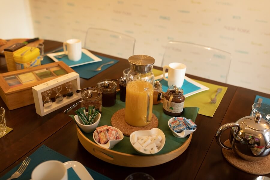 Focus on the center of the breakfast table with a large round wooden top. In the center, a glass bottle of orange juice and all around, jars of jams, spreads, small pieces of wrapped butter and sugar.