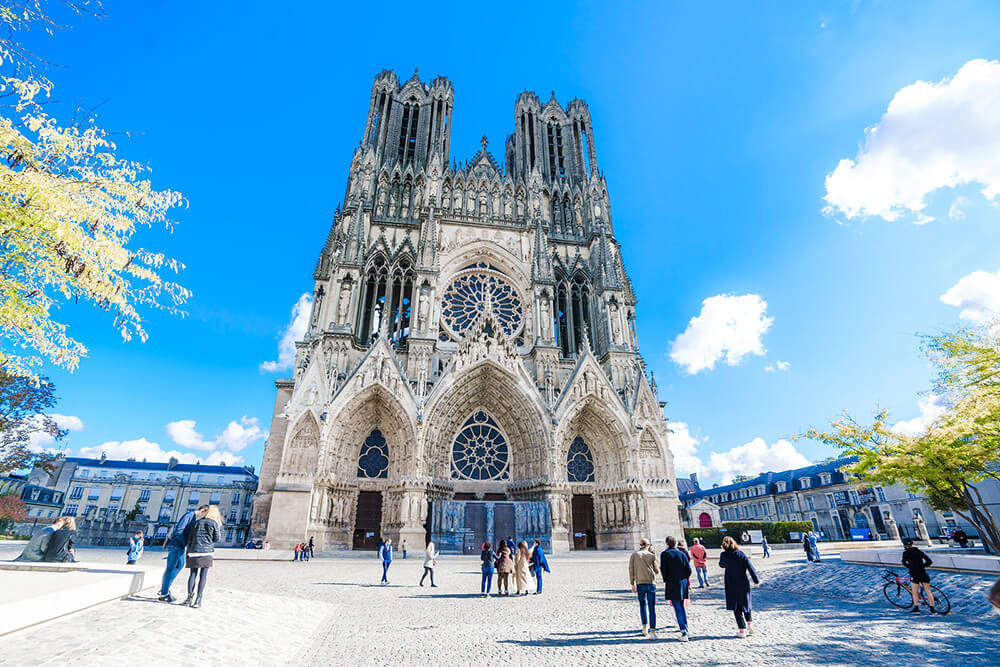 We see the majestic Reims Notre-Dame Cathedral, a masterpiece of Gothic architecture, and its square in front of a clear blue sky.